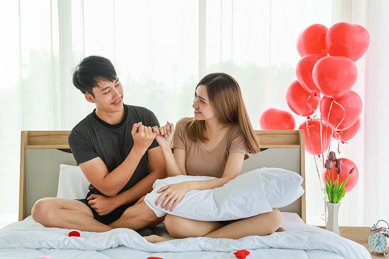 情侶節和情人節概念。亞洲男女在以紅色氣球裝飾的臥室的床上互相看著，手牽手。圖片下載