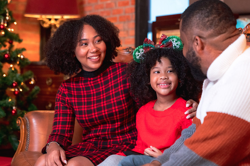 非裔美國小女孩在圣誕前夜和父母坐在裝飾好的客廳的地板上打開禮盒攝影圖片