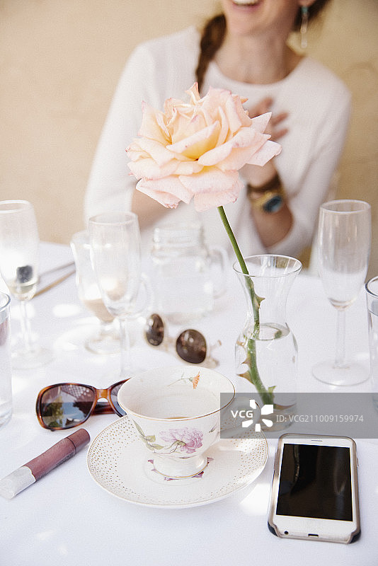 近距离的玫瑰花瓶在桌子上的杯子和玻璃杯，一个手机和太阳镜。图片素材