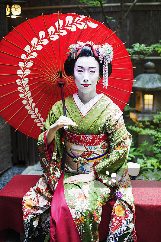 一名身穿传统艺妓风格的女子，穿着和服和宽腰带，梳着精致的发型和花发夹，化着白色的妆，鲜红色的嘴唇和黑色的眼睛，坐着撑着伞。图片素材