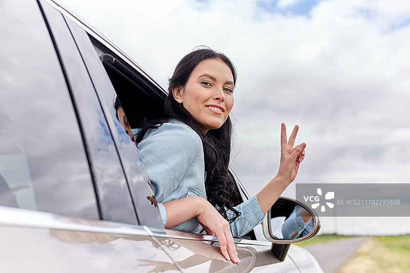暑假，假期，旅游，公路旅行和人们的概念-快乐的年轻女子开车和显示和平的标志。快乐的女人开着有和平标志的车图片素材