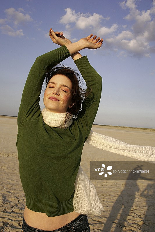 无忧无虑的女人穿着绿色毛衣，牛仔裤和围巾在海滩上享受阳光图片素材