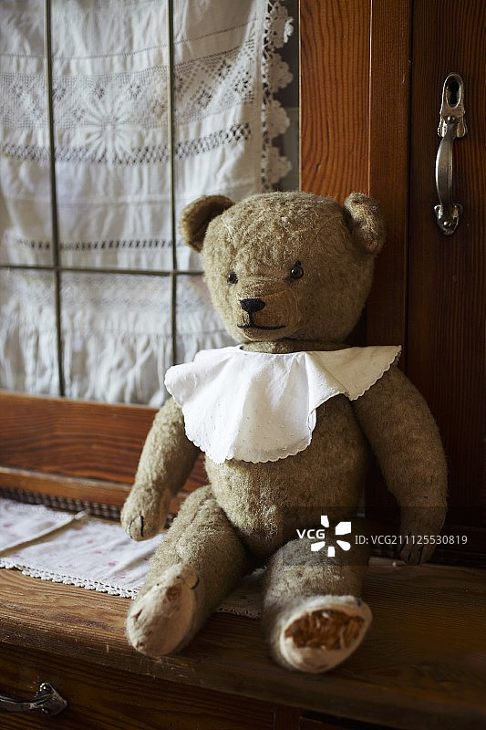 戴着围嘴的老泰迪熊坐在厨房梳妆台上，玻璃门上有花边窗帘图片素材