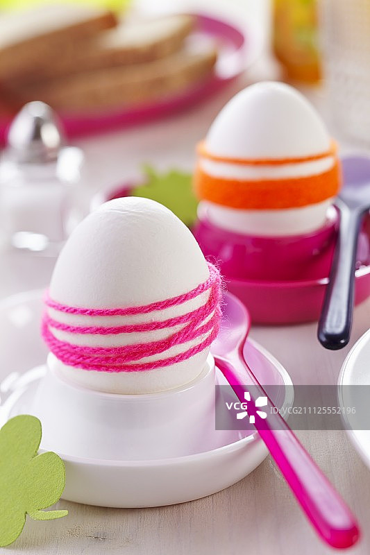 煮熟的鸡蛋用色彩鲜艳的毛线包裹在粉色和白色的蛋杯里，再配上塑料勺子图片素材