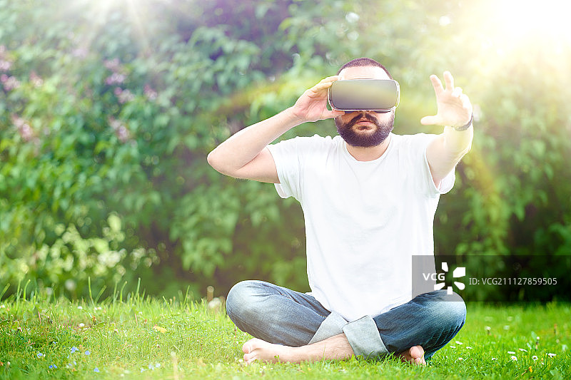 留着胡子的年轻男子戴着虚拟现实眼镜坐在花园里。生活方式VR的乐趣和…图片素材
