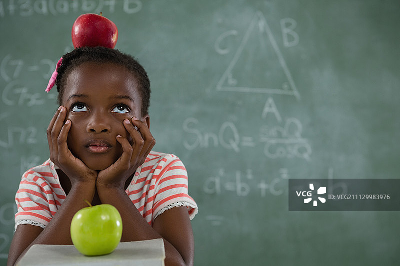 沉思的女学生坐在黑板前，头上戴着红苹果图片素材