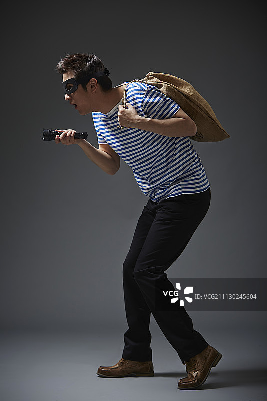 摄影棚拍摄的一个男人戴着黑色面具，穿着休闲服装，像小偷一样，肩上扛着一个袋子，在寻找带手电筒的东西图片素材