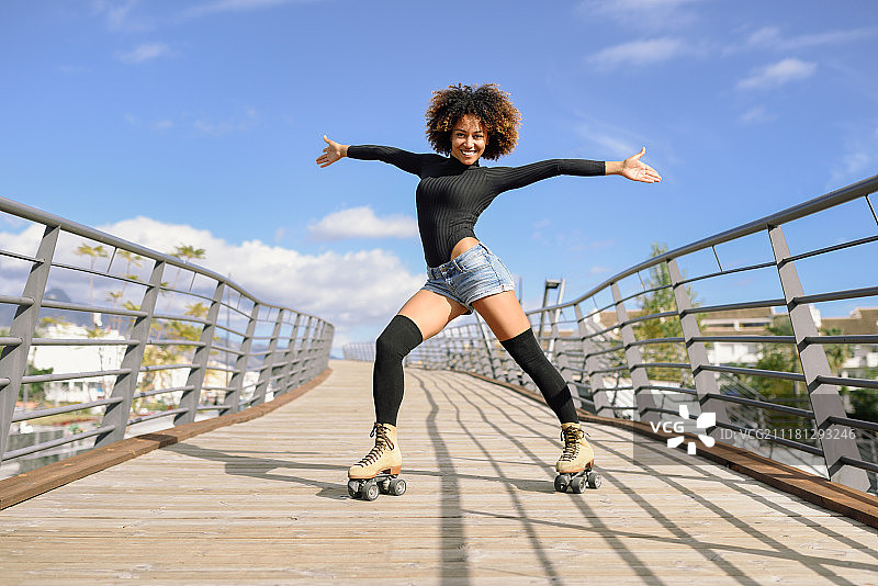 黑人妇女，非洲发式，溜冰鞋在户外城市桥上张开双臂。在阳光明媚的日子里，微笑着滑旱冰的年轻女性。天空中美丽的云彩。图片素材
