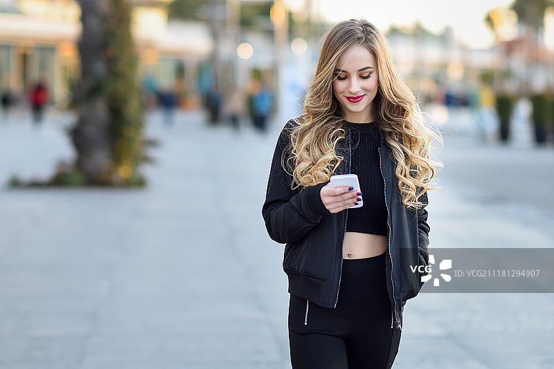 一个金发女人用她的智能手机在城市背景下发短信。金发女人用她的智能手机在城市背景下发短信。美丽的年轻女孩微笑着穿着黑色夹克走在街上。漂亮的俄罗斯女性与长波浪发型。图片素材