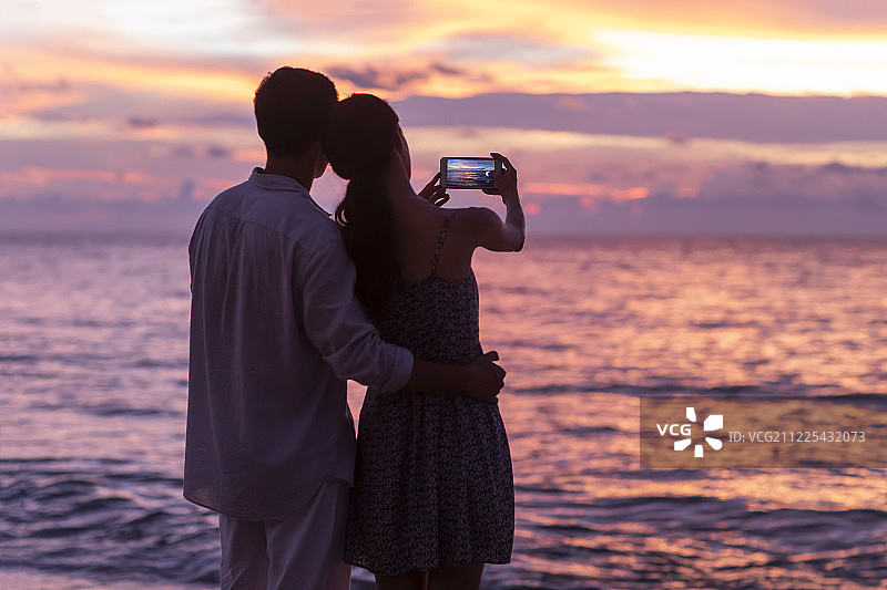 年轻情侣在海边使用手机拍摄海景图片素材