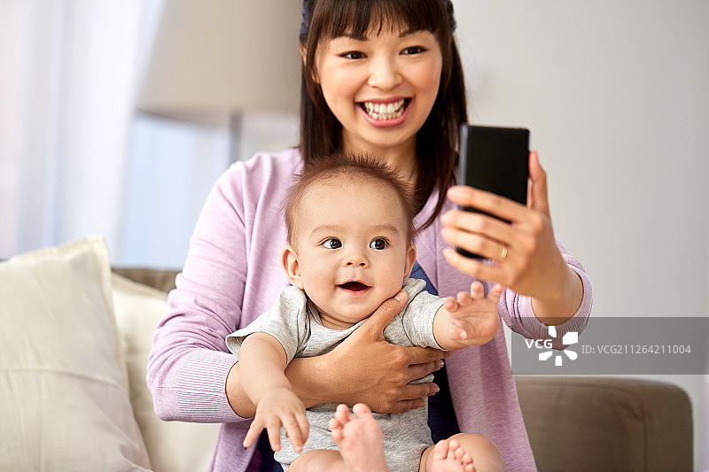 家庭和母亲的概念——快乐微笑的年轻亚洲母亲和小宝宝在家里用智能手机自拍。亚洲母亲和婴儿儿子在家里自拍图片素材