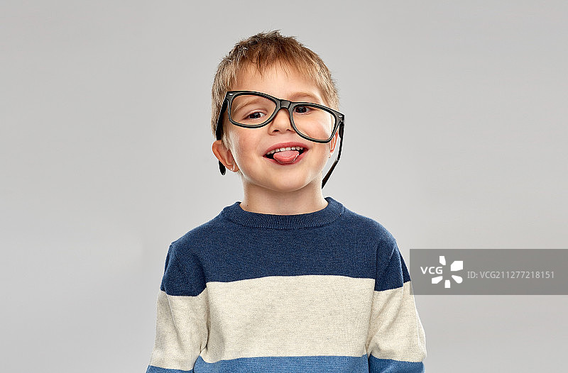 学校，教育和视觉概念-微笑的小男孩与弯曲戴眼镜显示舌头在灰色的背景。戴着眼镜露出舌头的小男孩的肖像图片素材