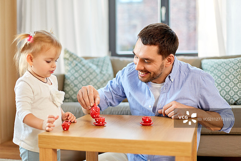 家庭，父亲和童年的概念——快乐的父亲和小女儿用玩具陶器在家里玩茶会。父亲和女儿在家里玩茶话会图片素材