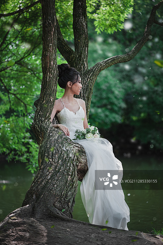 一名年轻女子穿着白色婚纱，手捧一束玫瑰，倚在公园的树上图片素材
