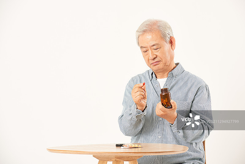 老人坐在桌边，手里拿着药瓶和药丸图片素材
