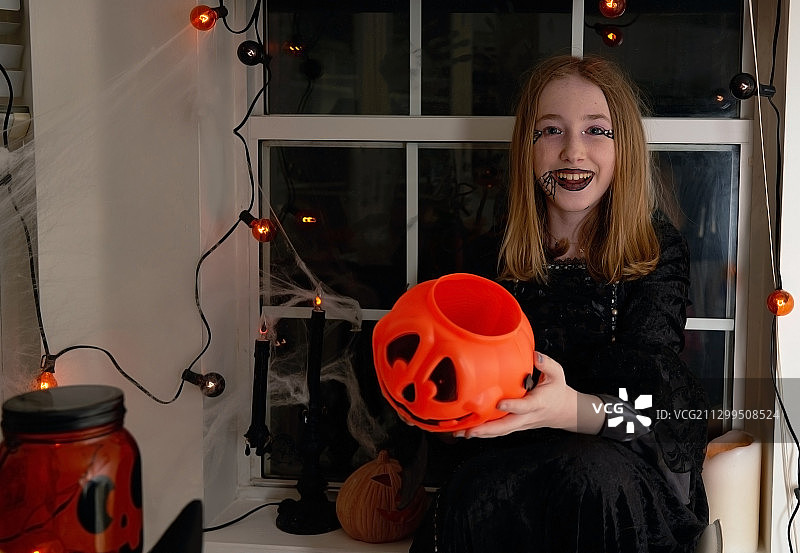 可爱的微笑女孩在万圣节装饰的房间里展示她的橙色杰克灯糖果桶图片素材