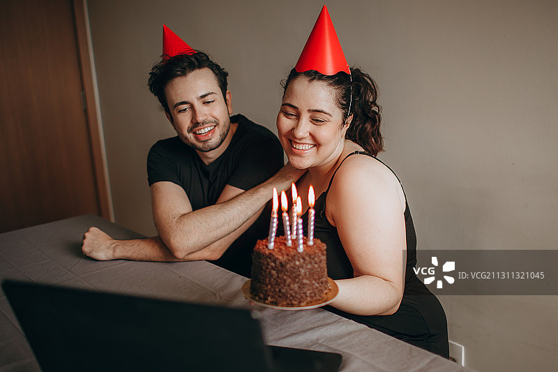 巴西米纳斯吉拉斯州贝洛奥里藏特，一对年轻的成年夫妇在家里用蛋糕和笔记本电脑庆祝生日图片素材