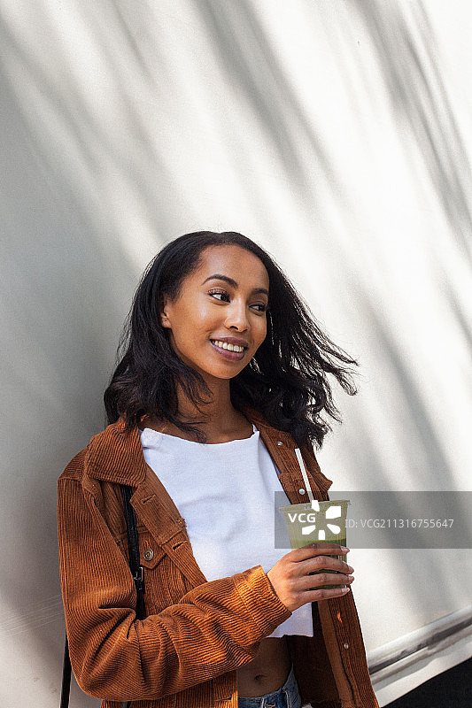 一个微笑的年轻女人拿着饮料，站在户外的墙，多伦多，安大略省，加拿大图片素材