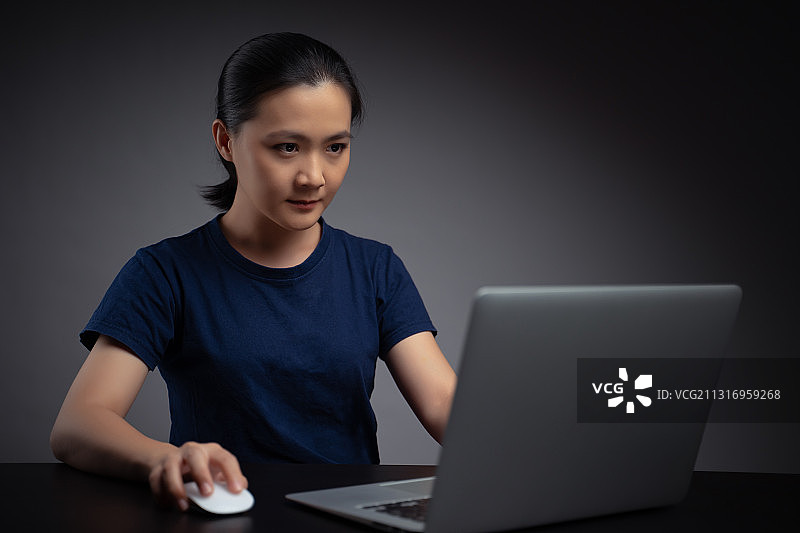 年轻女子坐在桌子前使用笔记本电脑，背景是灰色图片素材