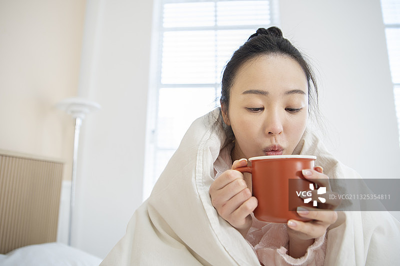 亚洲妇女在毛毯下穿着睡衣喝热茶，咖啡在马克杯图片素材