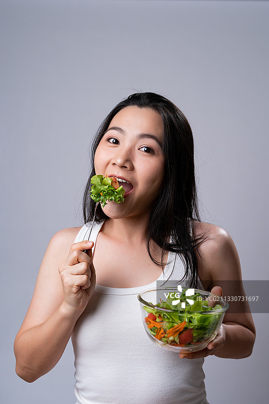 在灰色背景下，一个微笑的年轻女子吃沙拉的肖像图片素材