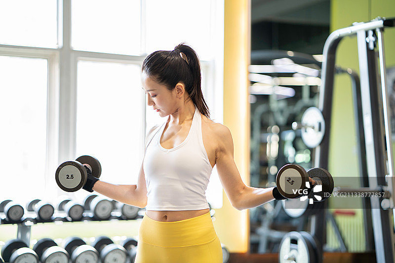 正在健身的年轻女性，在健身房使用健身器材哑铃锻炼。身材健美的年轻女人。图片素材