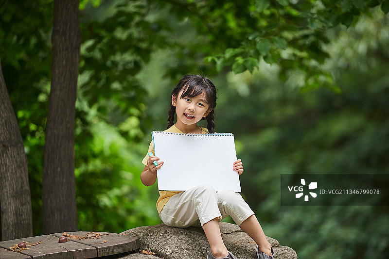 女孩坐在石头上，拿着空白的素描本，在森林里微笑图片素材