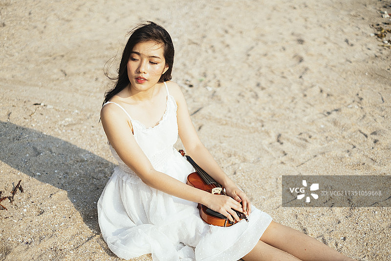黑色长发的小提琴手坐在沙滩上，小提琴放在她的屁股上。图片素材