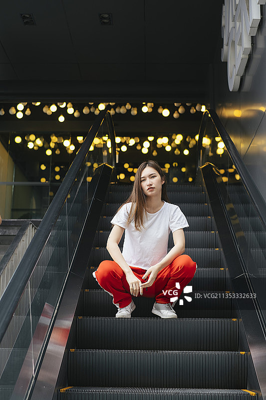 时尚肖像-美丽的亚洲年轻女子在白色衬衫和红色运动裤坐在自动扶梯上。后面是漂亮的散景背景。图片素材