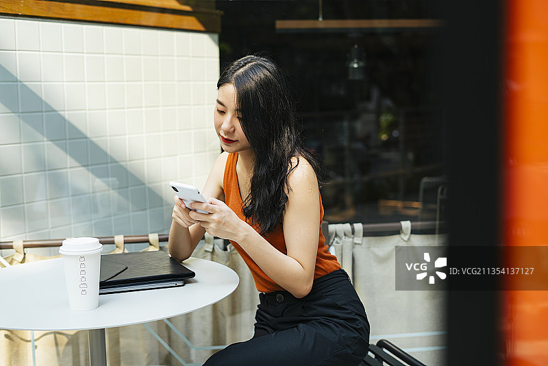 亚洲美女穿着橙色衬衫在咖啡店用智能手机。图片素材