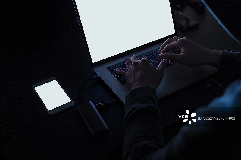 笔记本电脑屏幕上的拷贝空间-黑色连帽衫黑客在暗室用笔记本电脑入侵电脑网络。网络安全的概念。图片素材