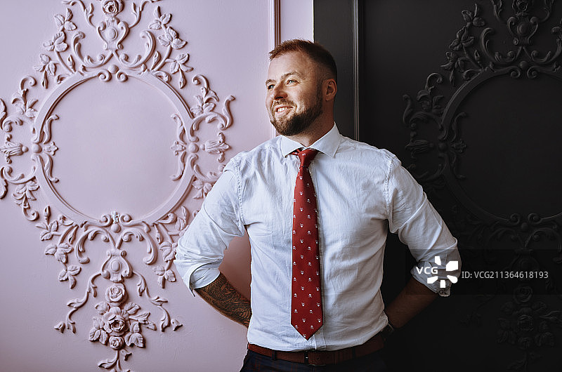 自信而成功的商人卷起白衬衫的袖子，系上红领带，在他办公室的黑粉色墙壁前摆姿势图片素材