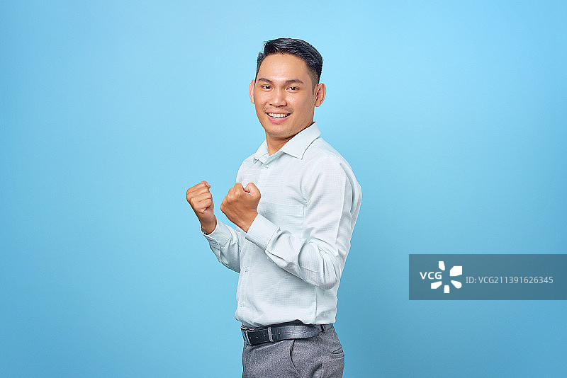 印度尼西亚，年轻英俊的商人在蓝色背景上举起手庆祝成功图片素材