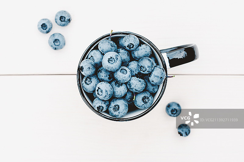 蓝莓-俯视杯满成熟的蓝莓，平放在白色的木桌图片素材