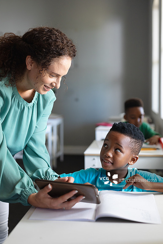 微笑的白人年轻女教师向非洲裔美国小学男孩展示数码平板电脑图片素材