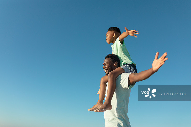 快乐的非洲裔美国男孩在父亲的肩膀上拷贝空间对晴朗的蓝天图片素材