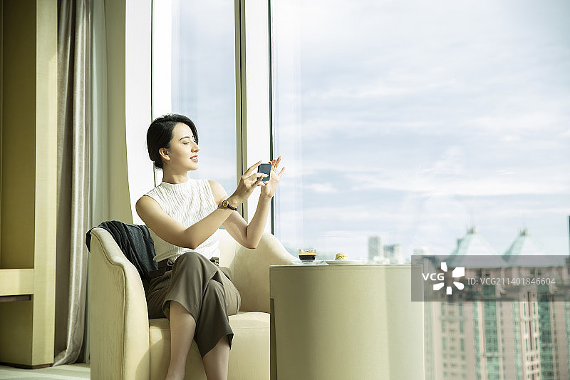 女商人在酒店房间休息喝下午茶，拿着手机拍窗外风景图片素材