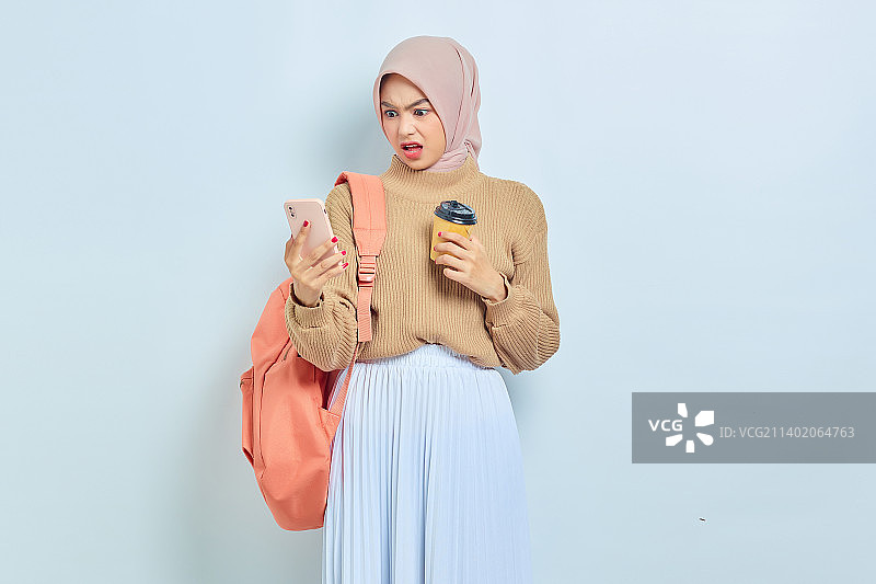 震惊的年轻亚裔穆斯林女学生，穿着棕色毛衣，拿着手机和咖啡图片素材