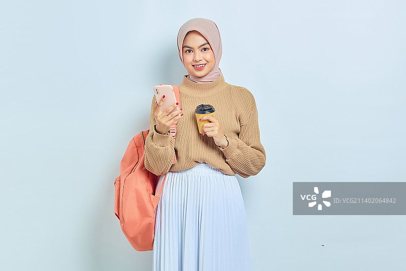 微笑的年轻亚裔穆斯林女学生，穿着棕色毛衣，拿着手机和咖啡图片素材