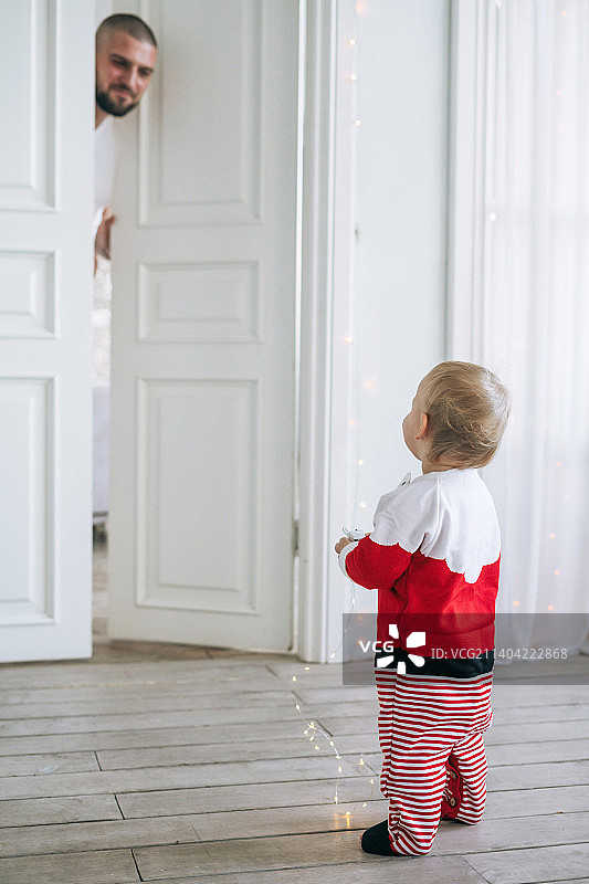 可爱的小女婴穿着圣诞老人的服装站在宽敞明亮的房间里看着爸爸，圣诞节的时候图片素材