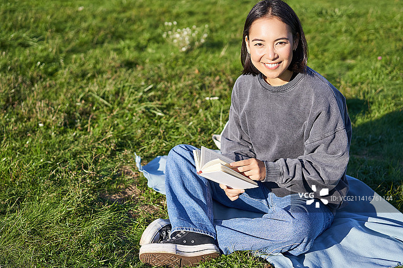 可爱的微笑的亚洲女孩坐在公园的毯子上，阅读和图片素材