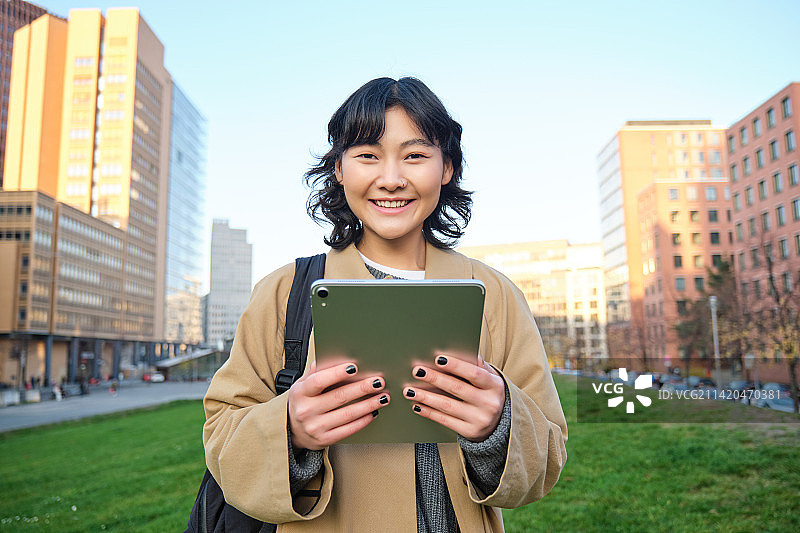 快乐的年轻黑发女孩，亚洲女人带着平板电脑在城市里散步图片素材