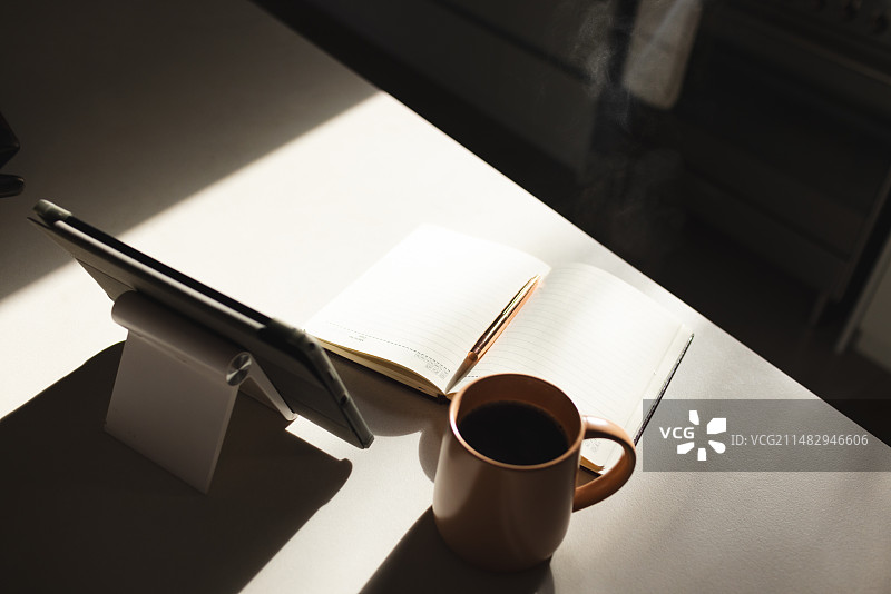 一个带触控笔的平板电脑，一个打开的笔记本，一杯咖啡放在一个有阳光的桌子上，上面有复印空间图片素材