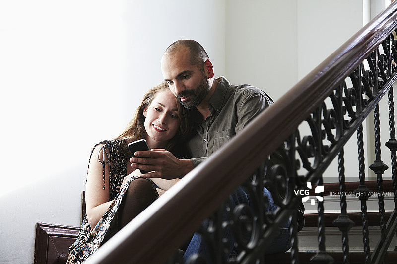一对年轻夫妇坐在楼梯上看手机短信的肖像。图片素材