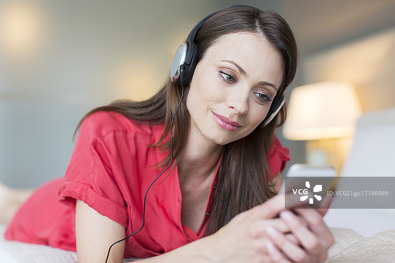 微笑的女人穿着红色裙子躺在床上听着智能手机上的音乐图片素材