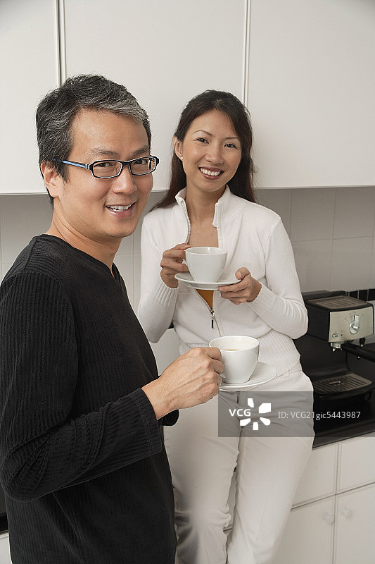 一个女人坐在厨房的柜台上，旁边站着一个男人，拿着咖啡杯，微笑着看着镜头。图片素材