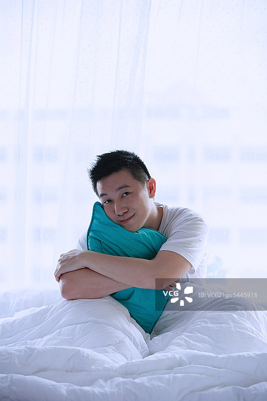 男人坐在床上，抱着枕头，对着镜头微笑图片素材