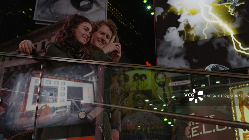 低角度的夫妇在阳台上拍摄标志性的广告牌广告在城市十字路口晚上图片素材