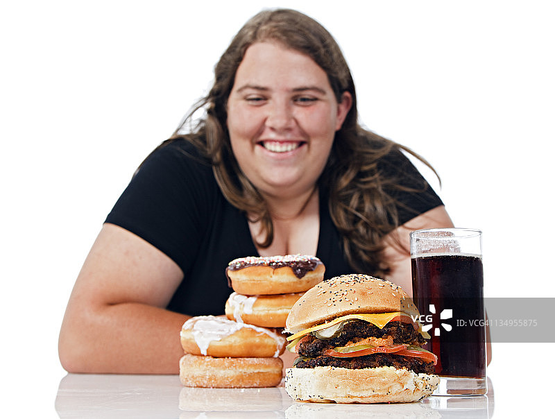 超重的年轻女子对着一堆让人发胖的垃圾食品微笑图片素材
