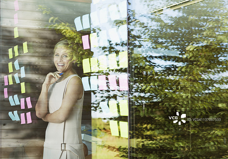 微笑的女商人看着办公室窗户上的不干胶便条的肖像图片素材